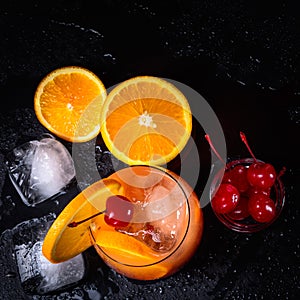 Tequila Sunrise cocktail, orange, ice cubes and maraschino cherries on a wet black slate tray. Top view with copy space.
