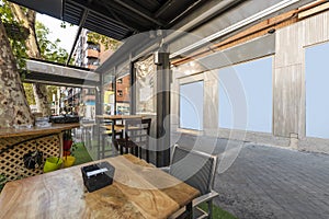 Terrace of a bar with artificial grass floor, wooden tables with gray armchairs and canvas and metal gazebo and polished metal