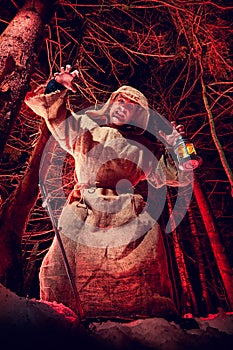 Terrible medieval monk in canvas sackcloth robe with lattern in dark forest and red light of moon on winter night. Fantasy or