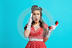 Terrified millennial woman in retro pinup dress holding landline phone receiver, touching her temples on blue background