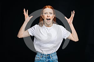 Terrified young woman in T-shirt shouting with hands at side on isolated back background