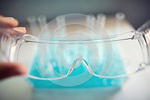 Test tubes are seen through protective goggles in a biology lab, hand holding transparent plastic glasses