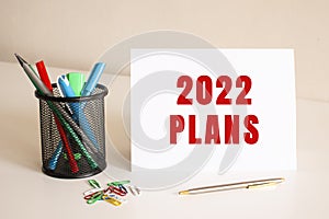 The text 2022 PLANS is written on a white folded sheet of paper on the table. Nearby are pens and pencils.