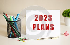 The text 2023 PLANS is written on a white folded sheet of paper on the table. Nearby are pens and pencils.