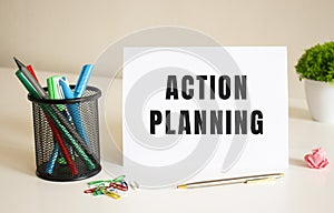 The text ACTION PLANNING is written on a white folded sheet of paper on the table. Nearby are pens and pencils.