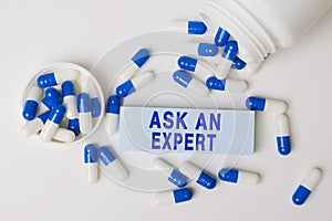Text - Ask an expert, written on paper note near blue-white pills spilling out of pill bottle. Medical concept