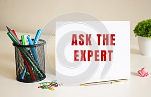 The text ASK THE EXPERT is written on a white folded sheet of paper on the table. Nearby are pens and pencils.