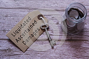 Text on brown tag with key and blurred jar full of coins on wooden table - Liquidated Ascertained Damages