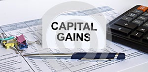 Text Capital Gains on white card with blue metal pen, calculator and paper clips on financial table