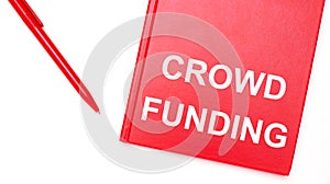 The text CROWD FUNDING is written on a red notepad near a red pen on a white table in the office. Business concept