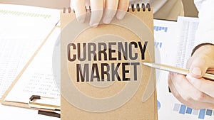 Text CURRENCY MARKET on brown paper notepad in businessman hands on the table with diagram. Business concept