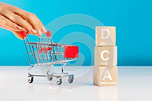 The text DCA is written on wooden cubes and woman's hand pushingn empty shopping cart in studio on blue background. Business