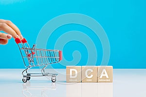 The text DCA is written on wooden cubes and womans hand pushingn empty shopping cart in studio on blue background. Business