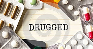 Text DRUGGED on a white background. Nearby are various medicines. Medical concept