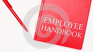 The text EMPLOYEE HANDBOOK is written on a red notepad near a red pen on a white table in the office. Business concept