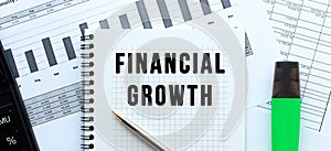 Text FINANCIAL GROWTH on the page of a notepad lying on financial charts on the office desk.