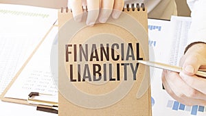 Text FINANCIAL LIABILITY on brown paper notepad in businessman hands on the table with diagram. Business concept