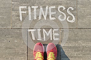 Text fitness time written on gray pavement with woman legs in sneakers, view from above