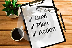 Text GOAL PLAN ACTION written in a clipboard next to a cup of coffee, eyeglasses and a green plant on a wooden table. Flat lay. To