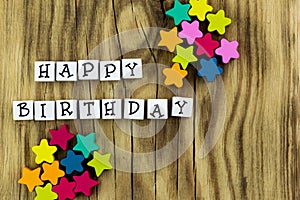 The text `happy birthday` arranged from blocks on a wooden background, next to colorful stars