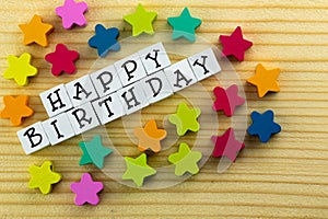 The text `happy birthday` arranged from blocks on a wooden background, next to colorful stars