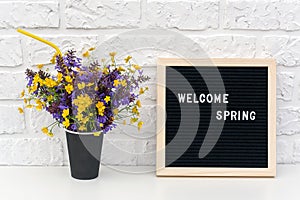Text Hello Spring on black letter board and bouquet of colored flowers in black paper coffee cup with cocktail straw on background