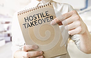 Text HOSTILE TAKEOVER on brown paper notepad in businessman hands in office. Business concept