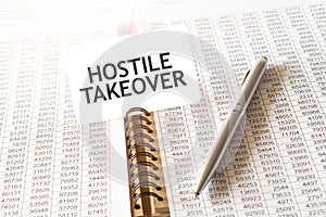 Text HOSTILE TAKEOVER on paper card, pen, financial documentation on table