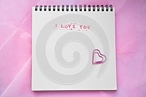 The text I love you in English, typed in stamps, on a white background, decorated with paper clips in the shape of a heart. Flat