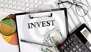 Text INVEST on Office desk table with keyboard, dollars,calculator ,supplies,analysis chart on the white background