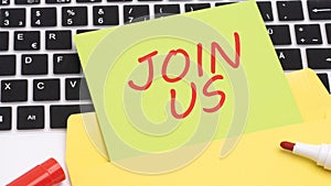 text JOIN US is written in red marker on a green card. in the frame is a laptop keyboard on yellow notepad. concept of