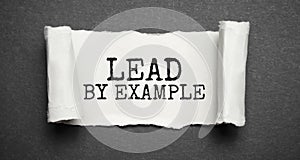 The text lead by example appearing behind torn grey paper