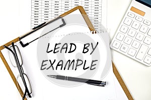 Text LEAD BY EXAMPLE on the white paper on clipboard with chart and calculator