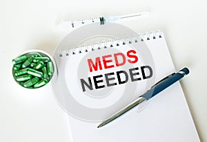 Text Meds Needed written in a notebook with a blue pen and a bunch of pills