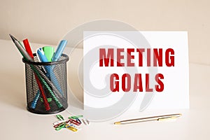 The text MEETING GOALS is written on a white folded sheet of paper on the table. Nearby are pens and pencils.