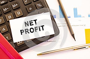 Text NET PROFIT on white card with metal pen, calculator and financial charts.