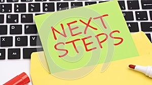 text NEXT STEPS is written in red marker on a green card. in the frame is a laptop keyboard on yellow notepad. concept