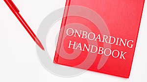 The text ONBOARDING HANDBOOK is written on a red notepad near a red pen on a white table in the office. Business concept