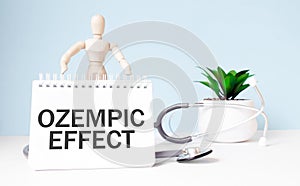 The text OZEMPIC EFFECT is written on notepad and wood man toy near a stethoscope on a blue background. Medical concept