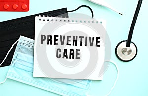 The text PREVENTIVE CARE is written in a notebook that lies on a blue background next to a thermometer and medical masks