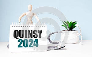 The text QUINSY 2024 is written on notepad and wood man toy near a stethoscope on a blue background. Medical concept