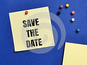 Text SAVE THE DATE written on yellow paper note with pin board over blue background.