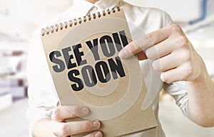 Text SEE YOU SOON on brown paper notepad in businessman hands in office. Business concept