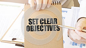 Text SET CLEAR OBJECTIVES on brown paper notepad in businessman hands on the table with diagram. Business concept