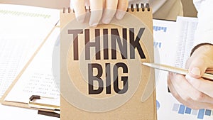 Text THINK BIG on brown paper notepad in businessman hands on the table with diagram. Business concept