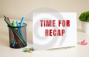 The text TIME FOR RECAP is written on a white folded sheet of paper on the table. Nearby are pens and pencils.
