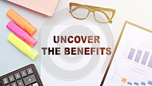 The text - UNCOVER THE BENEFITS on office desk with calculator, markers, glasses and financial charts
