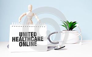 The text UNITED HEALTHCARE ONLINE is written on notepad and wood man toy near a stethoscope on a blue background. Medical concept