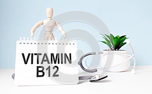 The text VITAMIN B12 is written on notepad and wood man toy near a stethoscope on a blue background. Medical concept