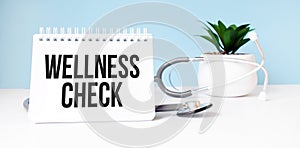 The text wellness check is written on notepad near a stethoscope on a blue background. Medical concept